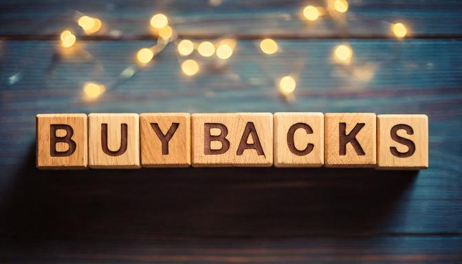Wooden letter tiles spelling the word BUYBACKS on a wooden surface with warm bokeh lights behind