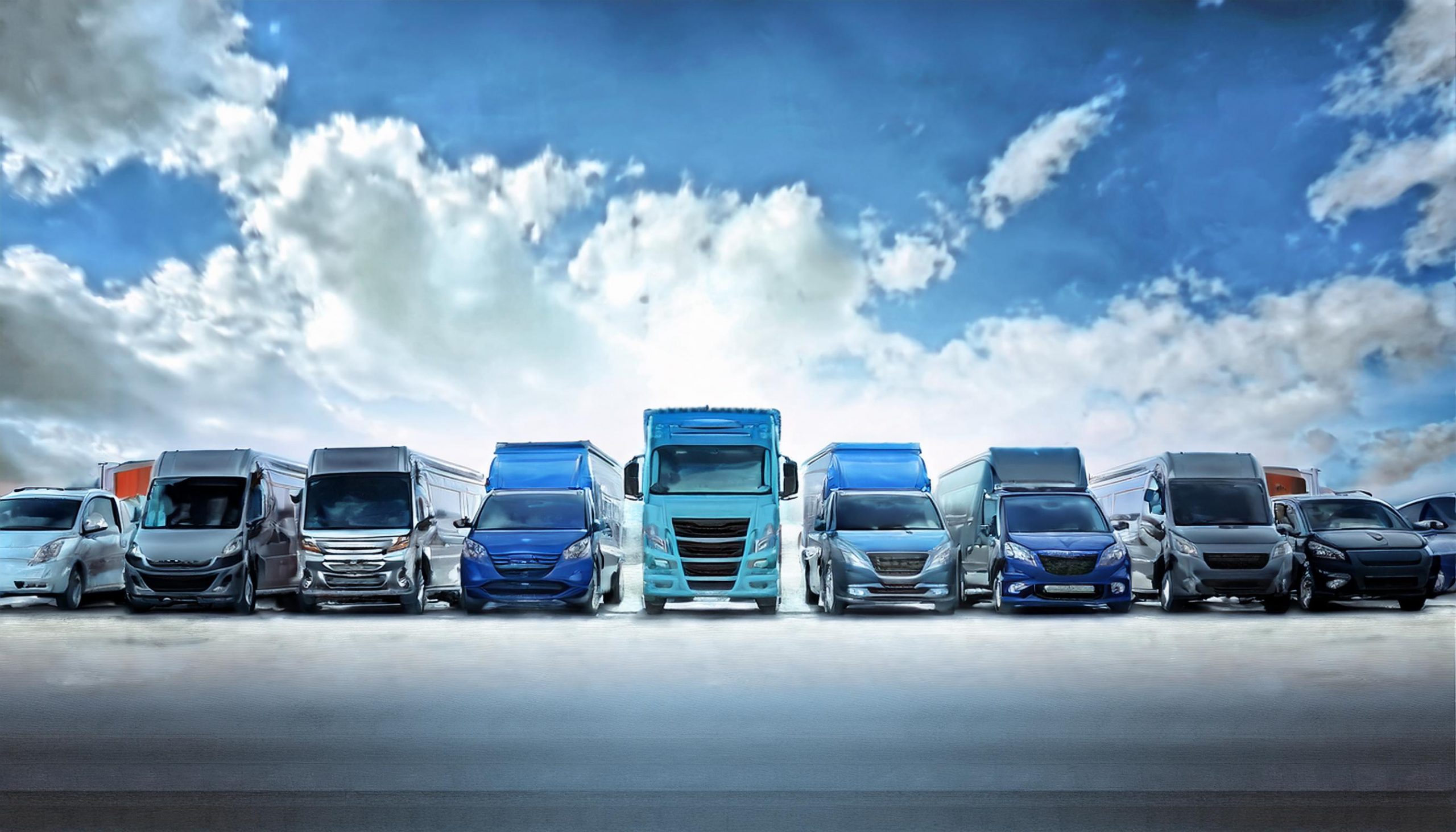 A fleet of vans, trucks and cars lined up under a cloudy blue sky, with a large cyan truck at the centre